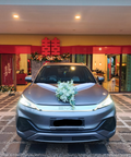 A metallic gray sedan parked facing forward in a driveway or entrance area, decorated with a white floral bouquet on the hood. The background shows a decorated venue with red banners, Chinese double happiness symbols, and warm interior lighting.