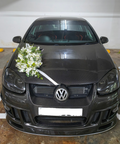 A dark gray Volkswagen hatchback car parked in a garage, decorated with a white floral bouquet on the hood and a white ribbon across the front grille.