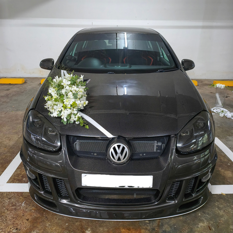 A dark gray Volkswagen hatchback car parked in a garage, decorated with a white floral bouquet on the hood and a white ribbon across the front grille.