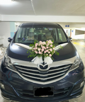 A dark blue minivan parked in an indoor parking structure, decorated with a pink and white floral arrangement and white ribbons across the hood. The front grille features a chrome Mazda logo, and the license plate reads SNK 9365P.
