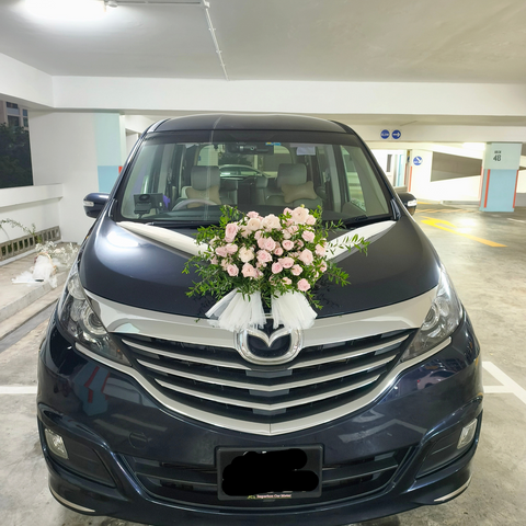 A dark blue minivan parked in an indoor parking structure, decorated with a pink and white floral arrangement and white ribbons across the hood. The front grille features a chrome Mazda logo, and the license plate reads SNK 9365P.