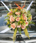 A decorative floral arrangement with blush pink roses, green foliage, and orange tropical leaves, tied to the windshield of a black car.