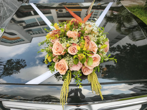 A decorative floral arrangement with blush pink roses, green foliage, and orange tropical leaves, tied to the windshield of a black car.