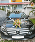 A black Mercedes-Benz sedan parked outdoors in front of a building, decorated with white ribbons and a floral arrangement on the hood. The front license plate reads SNY 5541A.