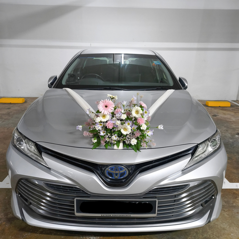 A silver Toyota sedan parked indoors, decorated with a pink and white floral arrangement on the hood and a white ribbon bow. The front license plate reads SJF 9811S.