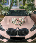 A light pink BMW sedan parked outdoors on a paved driveway, decorated with a white and pink floral arrangement on the hood. The front grille and license plate are visible, with another pink car partially visible on the right.