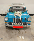 A classic blue wedding car with a white roof, decorated with a large white floral arrangement on the hood, white ribbons across the front, and a chrome bumper. The license plate reads SNM 9008 K.