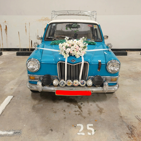 A classic blue wedding car with a white roof, decorated with a large white floral arrangement on the hood, white ribbons across the front, and a chrome bumper. The license plate reads SNM 9008 K.