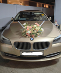 A gold BMW sedan photographed from the front in a garage or showroom. The car is decorated with a large floral arrangement on the hood and white ribbons across the windshield.