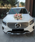 A white Mercedes-Benz SUV parked outdoors on a paved driveway, decorated with a pink and white floral arrangement on the hood. The front grille features the Mercedes-Benz logo, and the license plate reads SNZ6534 J.