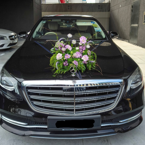 A black Mercedes-Benz sedan photographed from the front in a driveway or parking area. The hood is decorated with a floral arrangement in pink, purple, and green, and the Mercedes-Benz emblem is visible at the center of the grille.