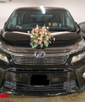 A black Hyundai minivan parked indoors, decorated with a pink and white floral arrangement on the hood. The front grille, headlights, and license plate are visible, with another dark vehicle partially visible on the right.
