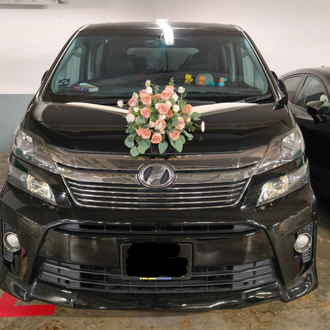 A black Hyundai minivan parked indoors, decorated with a pink and white floral arrangement on the hood. The front grille, headlights, and license plate are visible, with another dark vehicle partially visible on the right.
