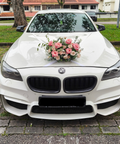 A white BMW sedan parked outdoors on a paved driveway, decorated with a pink and peach floral arrangement on the hood. The front grille, headlights, and license plate are visible, with grass, trees, and buildings in the background.