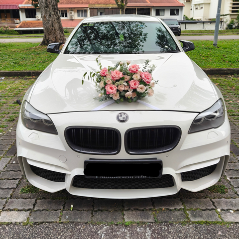 A white BMW sedan parked outdoors on a paved driveway, decorated with a pink and peach floral arrangement on the hood. The front grille, headlights, and license plate are visible, with grass, trees, and buildings in the background.