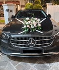 A black Mercedes-Benz sedan parked outdoors on a stone driveway, decorated with white ribbons across the hood and a pink and white floral arrangement on the front grille.