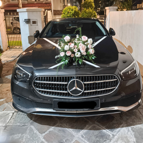 A black Mercedes-Benz sedan parked outdoors on a stone driveway, decorated with white ribbons across the hood and a pink and white floral arrangement on the front grille.