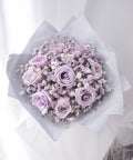 Lilac roses and purple baby's breath in pale grey wrapping beside a curtain
